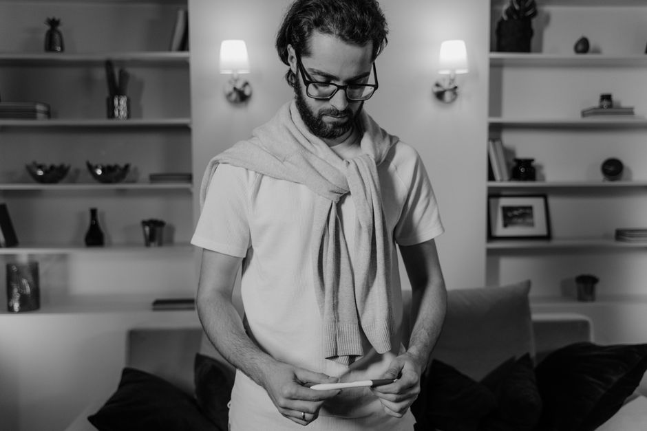 A man in glasses holding a pregnancy test, standing in a cozy room.
