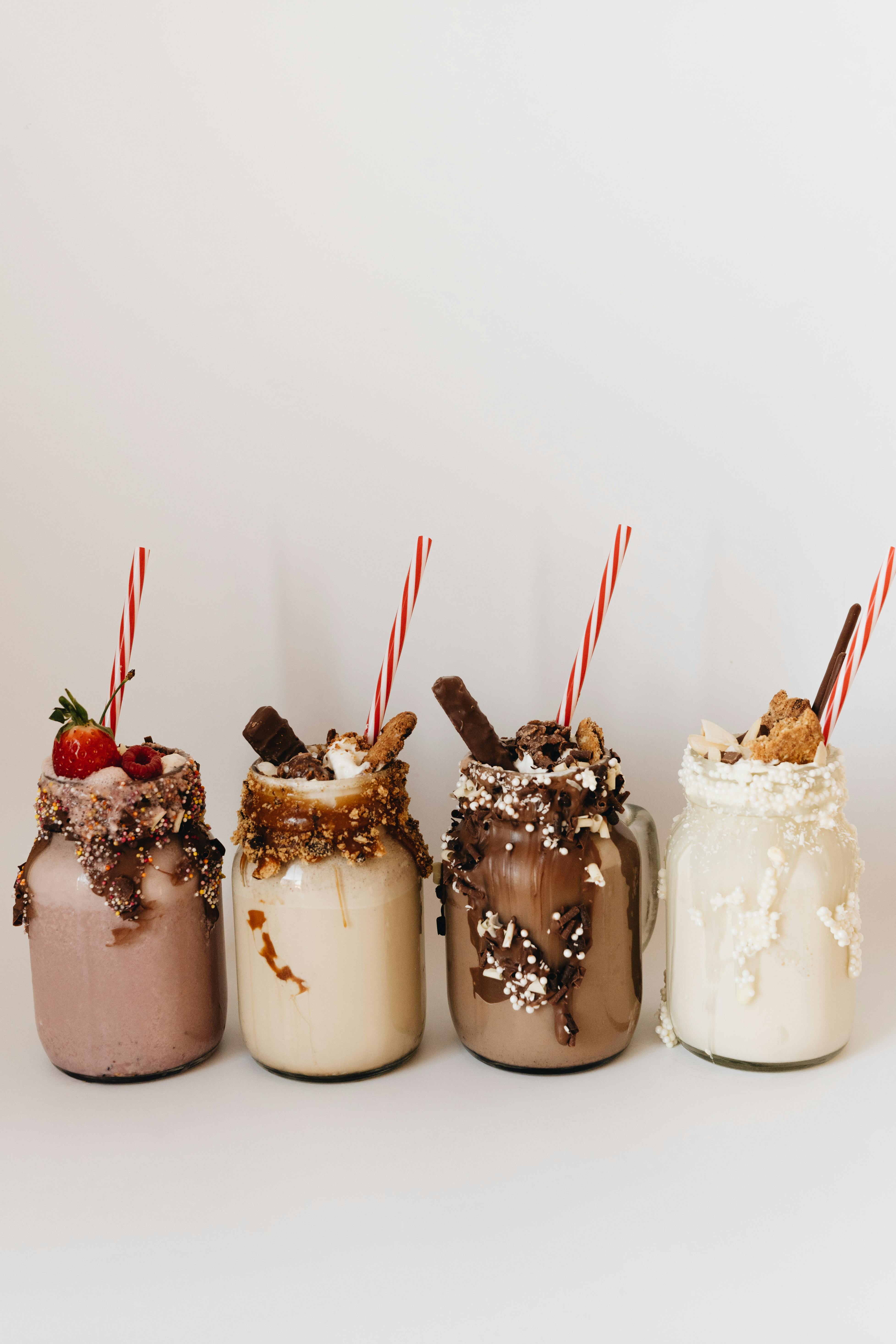 Four gourmet milkshakes in mason jars with various toppings and straws on a white background.
