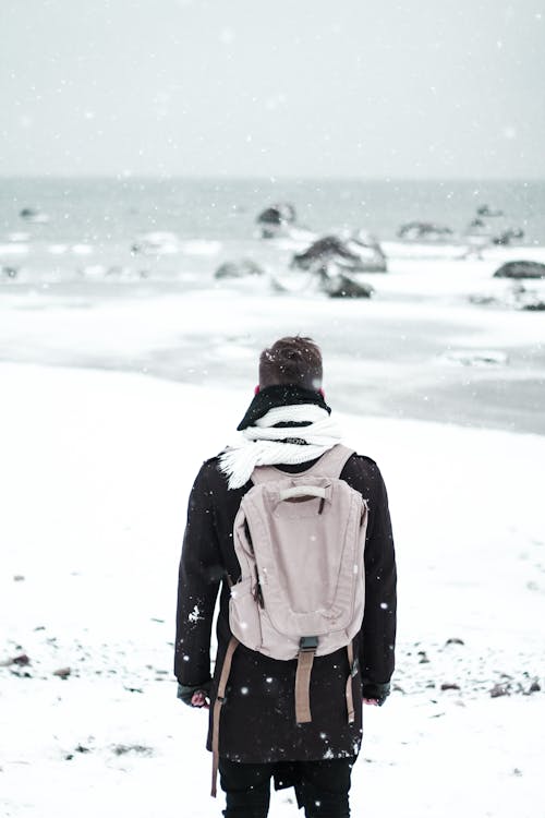 Back view of unrecognizable male in outerwear with rucksack standing on snowy coast near water with stones in winter time