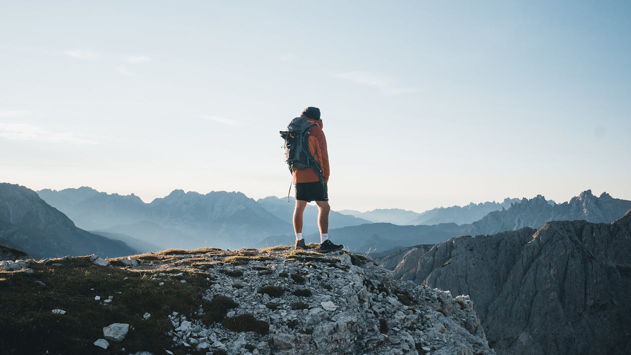 Free Back view anonymous male explorer with backpack standing on rough mountain peak and enjoying breathtaking scenery of severe highland on clear day Stock Photo