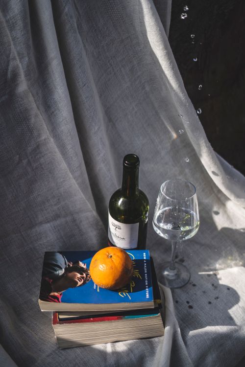 Free A stylish still life composition featuring wine, books, and an orange against a fabric backdrop. Stock Photo