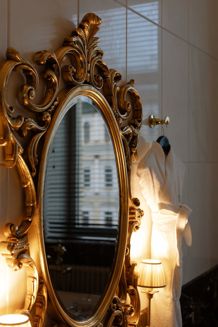 Classic Oval Mirror Hanging On Wall Beside Two Lighted Lamps And Bathrobe