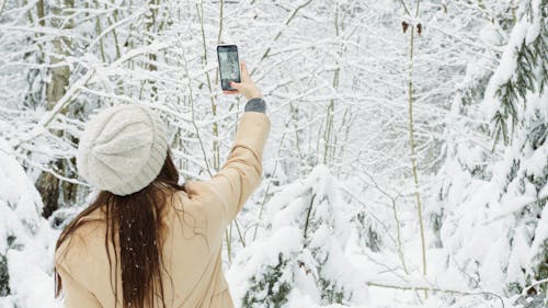 Woman in White Knit Hat Taking Photo of Snow Covered Trees