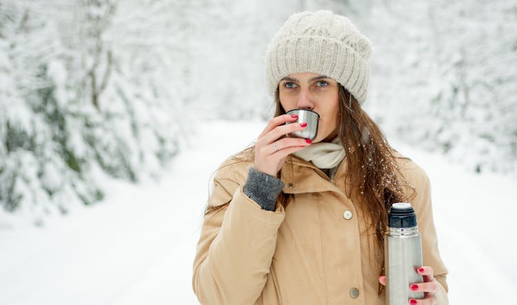 Woman In Beige Coat Standing On Snow Covered Ground Drinking From A Thermos