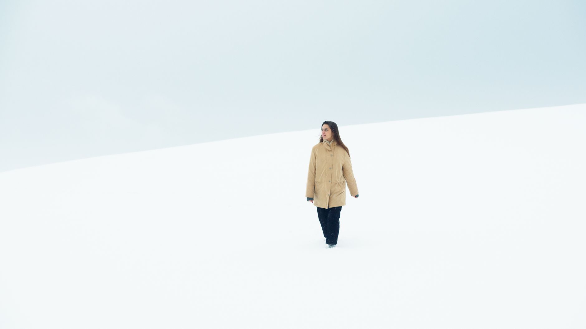 Woman strolling alone in a vast snowy field, wearing a brown coat during winter.