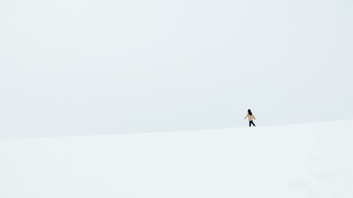 Free A woman walks alone in a vast, snowy landscape under a minimalistic, pale sky. Stock Photo
