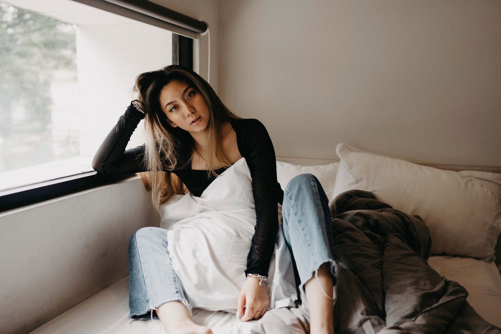 Woman in a black top and jeans relaxing on a bed beside a bright window