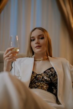 Chic woman in white blazer, holding a wine glass indoors, exuding elegance.