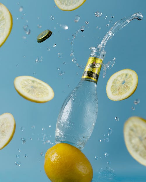 Creative composition of opened bottle with refreshing soft drink soaring on blue background near ripe lemon slices