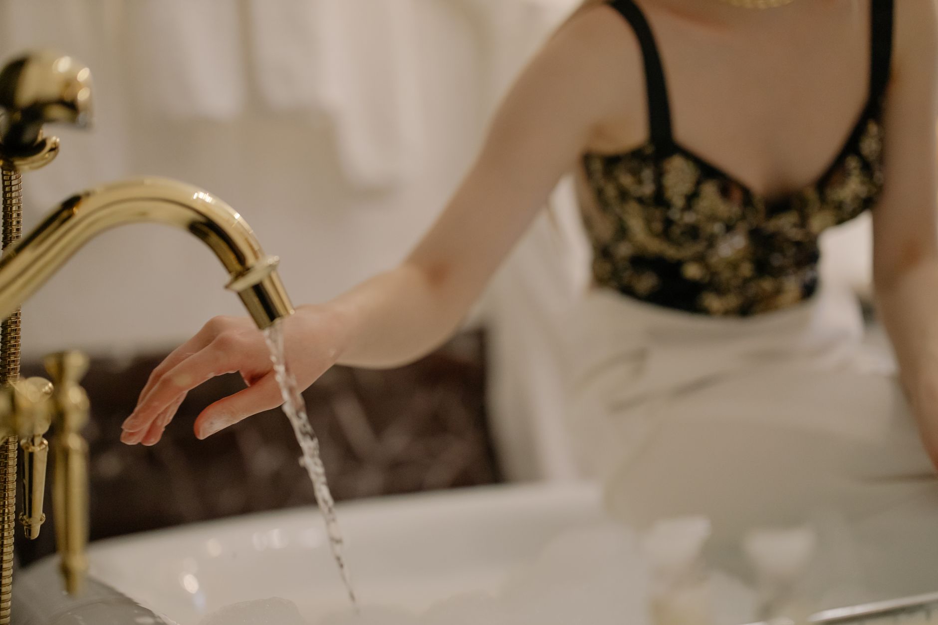 An elegant woman washes her hand under a golden faucet indoors.