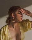 Feminine young lady in trendy dress with low neckline and elegant earrings covering eyes with hand while looking on bright light in studio