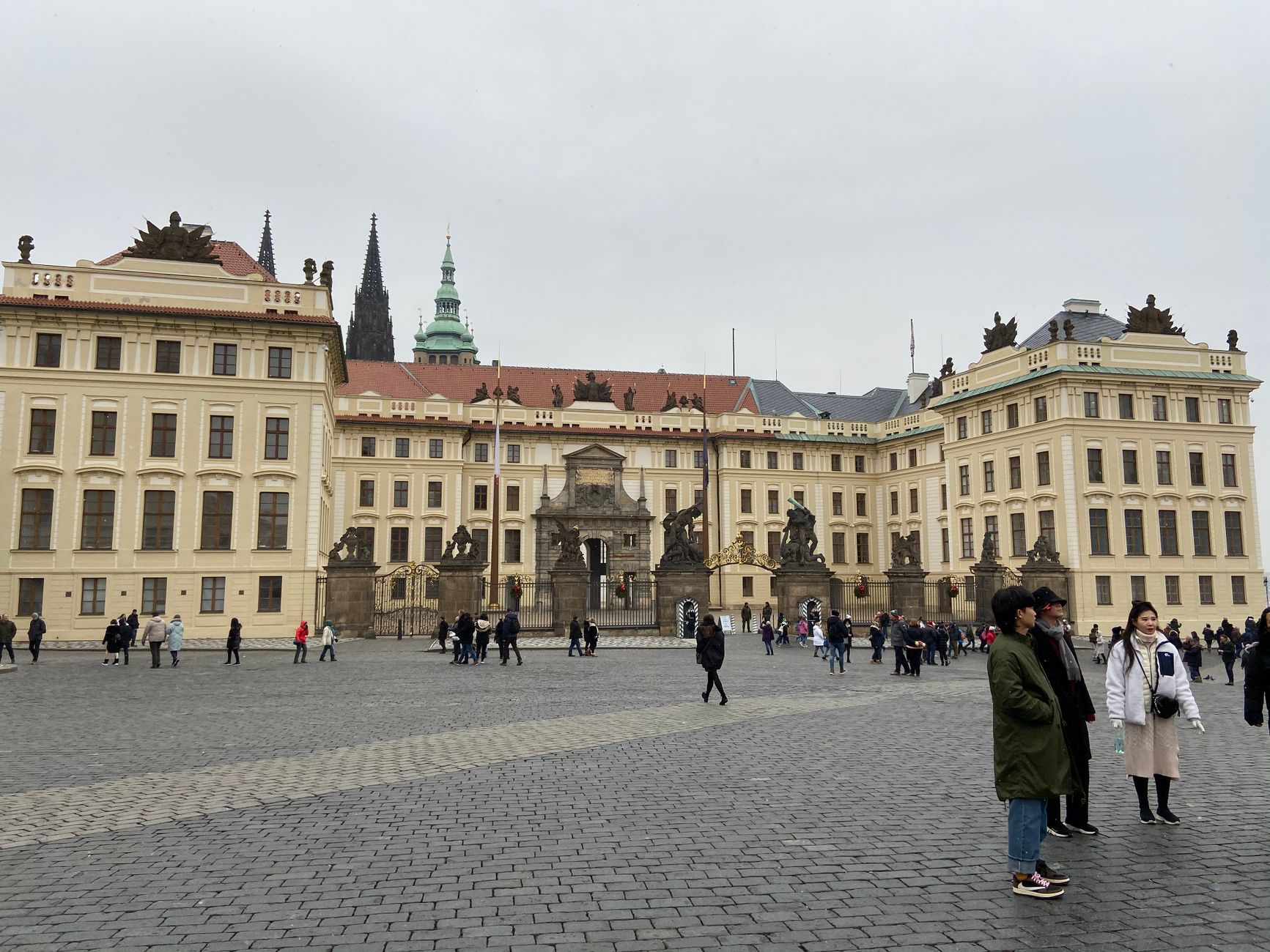 Tourists explore Prague Castle's grand facade under a gray, cloudy sky.