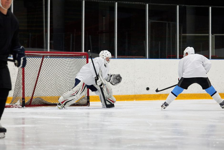 Ice hockey action scene featuring a goalie in white gear defending against an approaching player indoors.