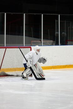 A hockey goalie in white gear stands ready to defend the goal in an indoor ice rink.