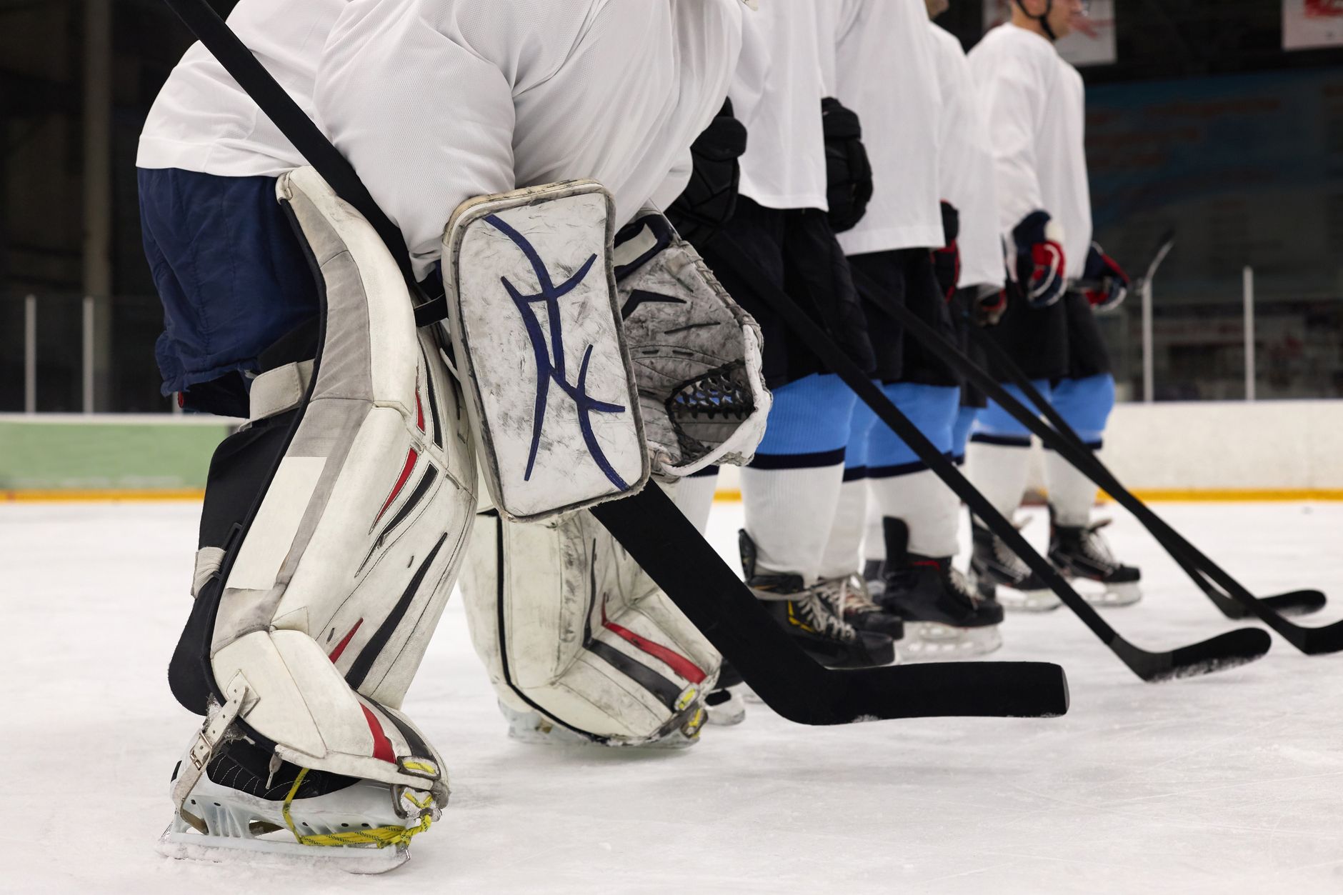 Close-up of ice hockey players in formation with goalie pads, sticks, and uniforms on a rink.