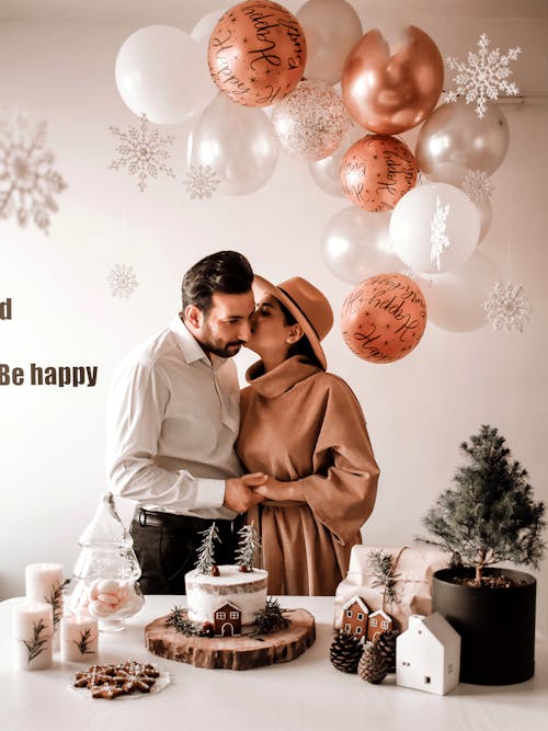 Woman hugging and kissing man while congratulating with birthday on surprise party with small Christmas tree and homemade cake on table against balloons