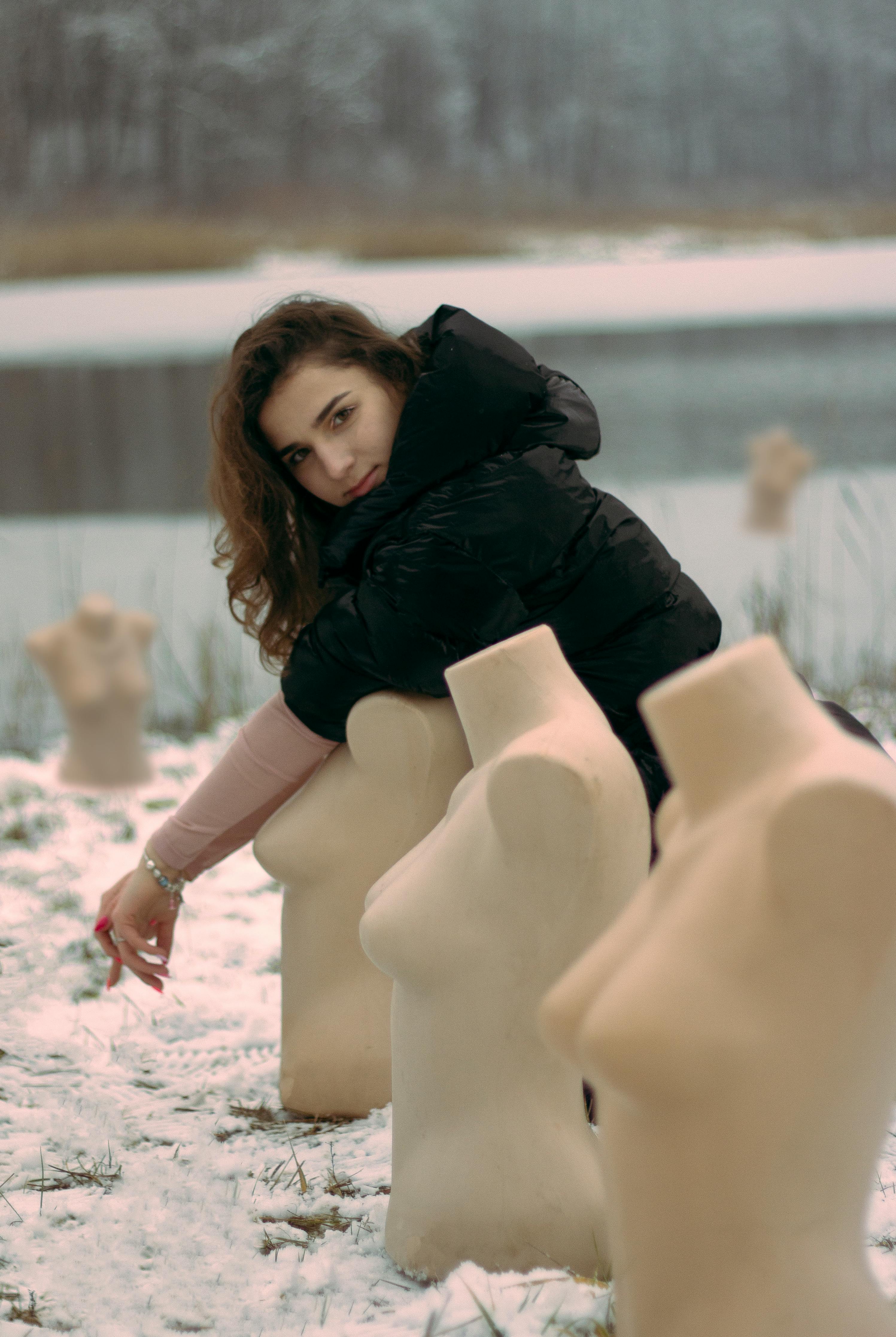 A woman in a hoodie jacket poses with mannequins by a snowy lakeside, creating a whimsical winter scene.