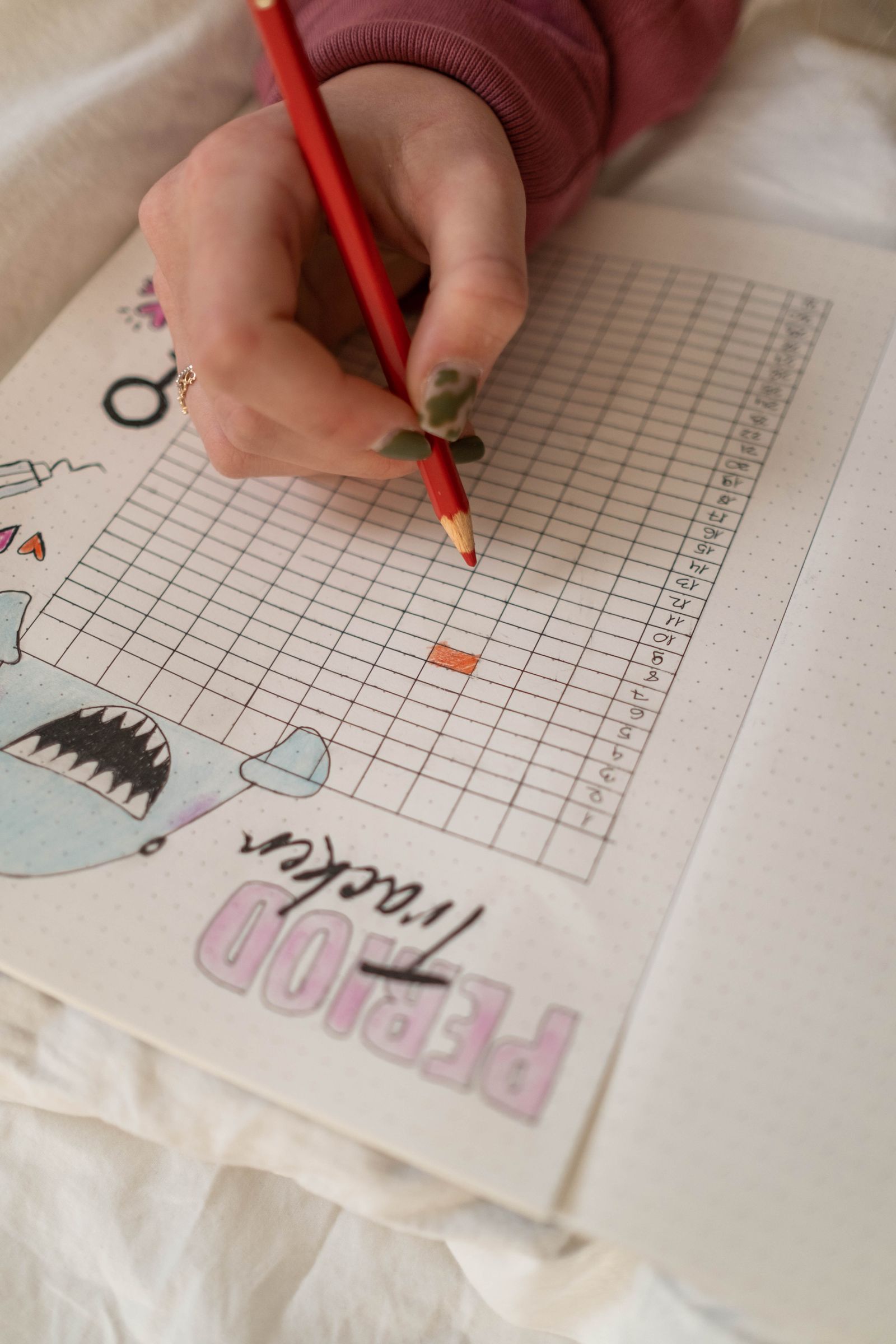 A hand marking cycle days on a paper period tracker in a notebook with a red colored pencil