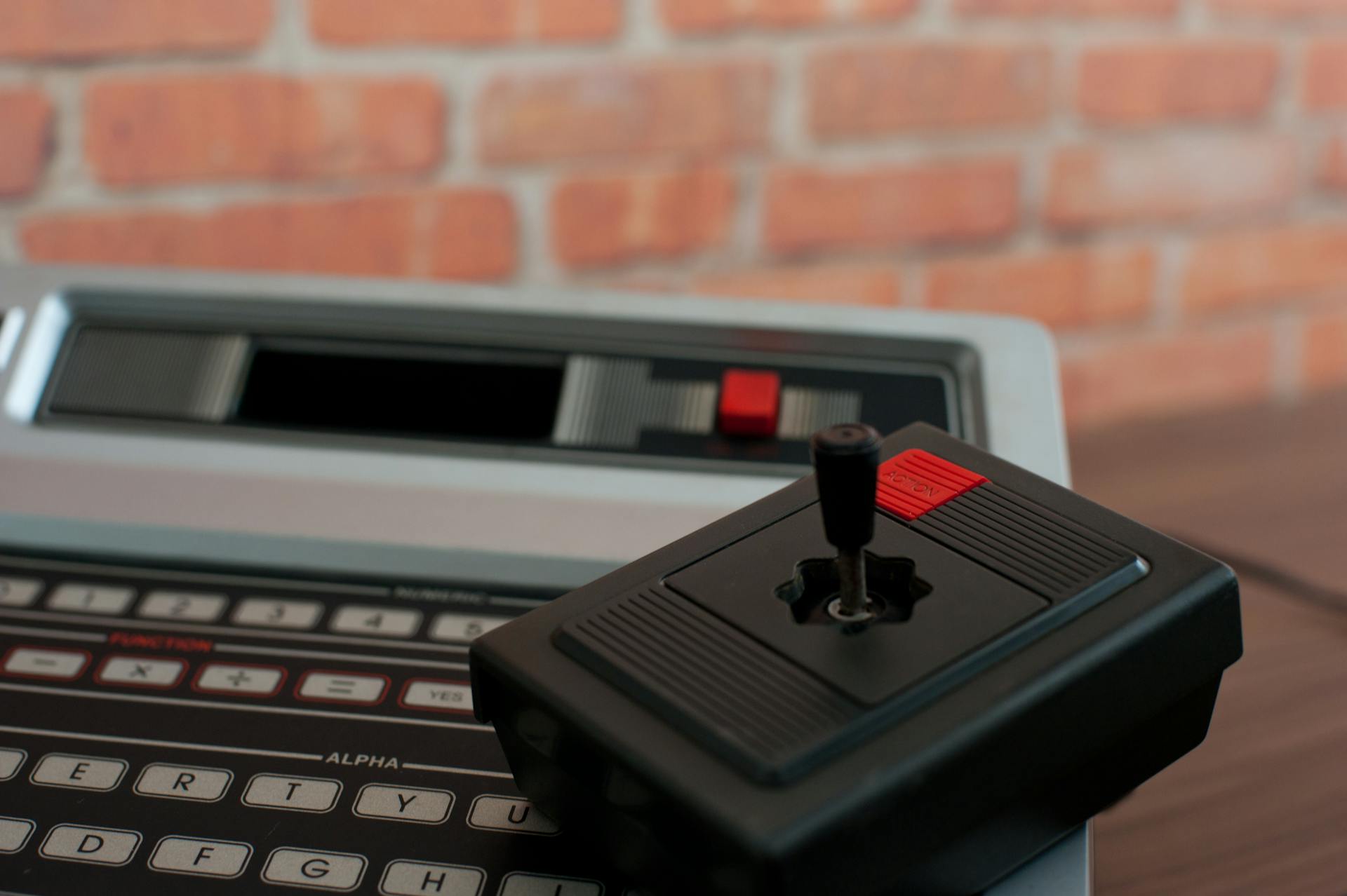 A close-up of a vintage Philips Odyssey 2 game console and joystick against a brick wall.