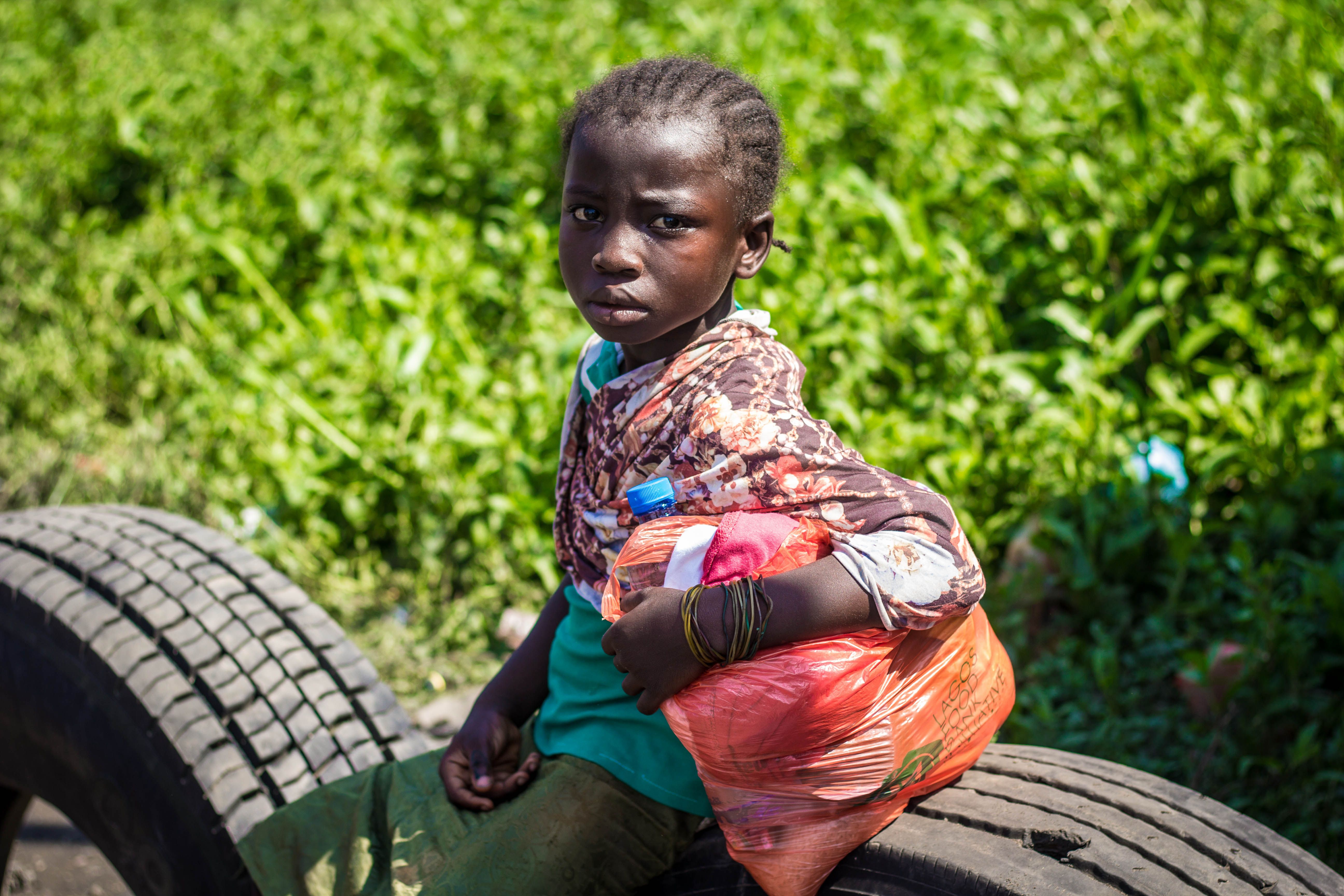African child holding a plastic bag, sitting on a tire outdoors in bright light.
