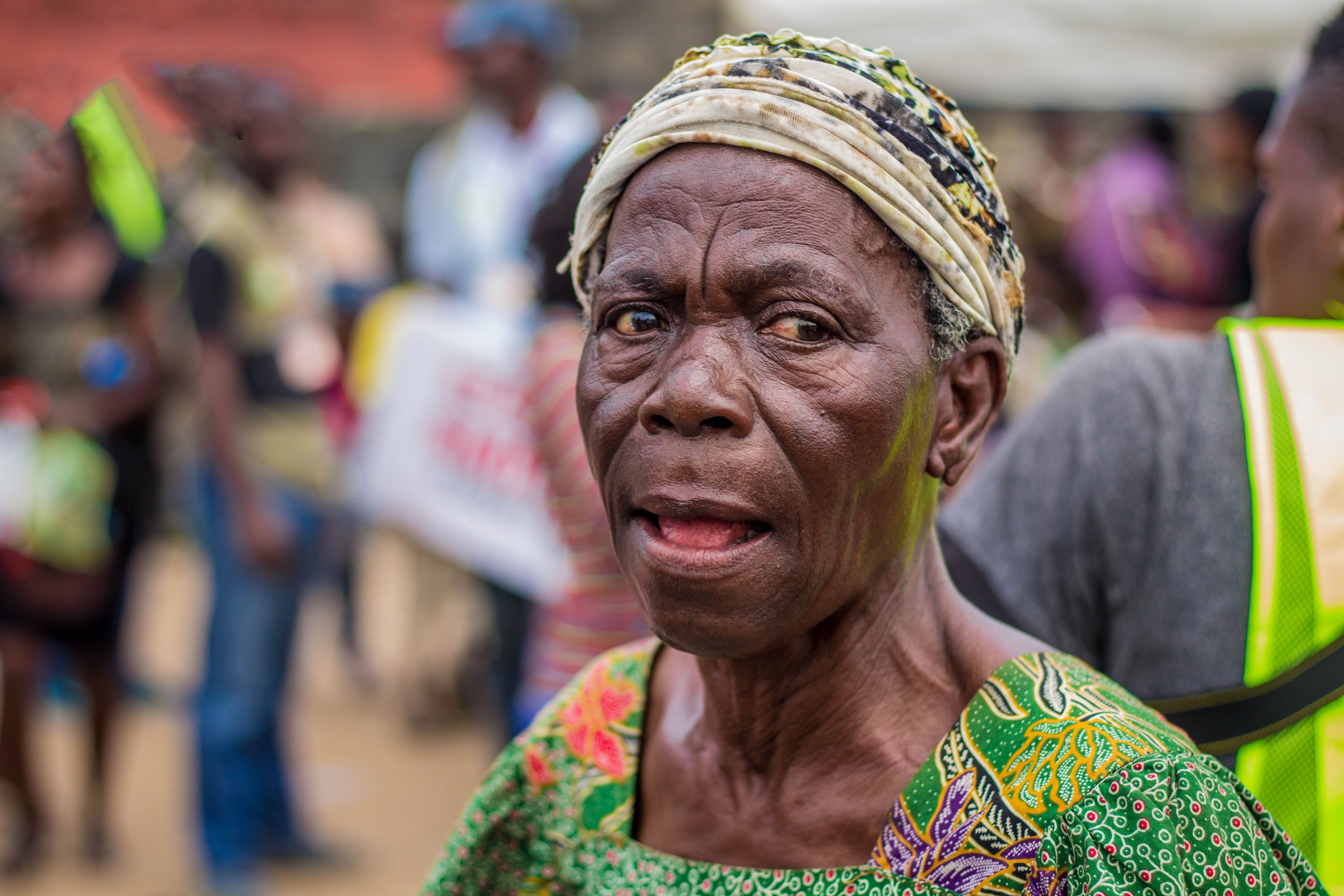 Close-up portrait of an elderly African woman wearing a headscarf, outdoors in a vibrant setting.