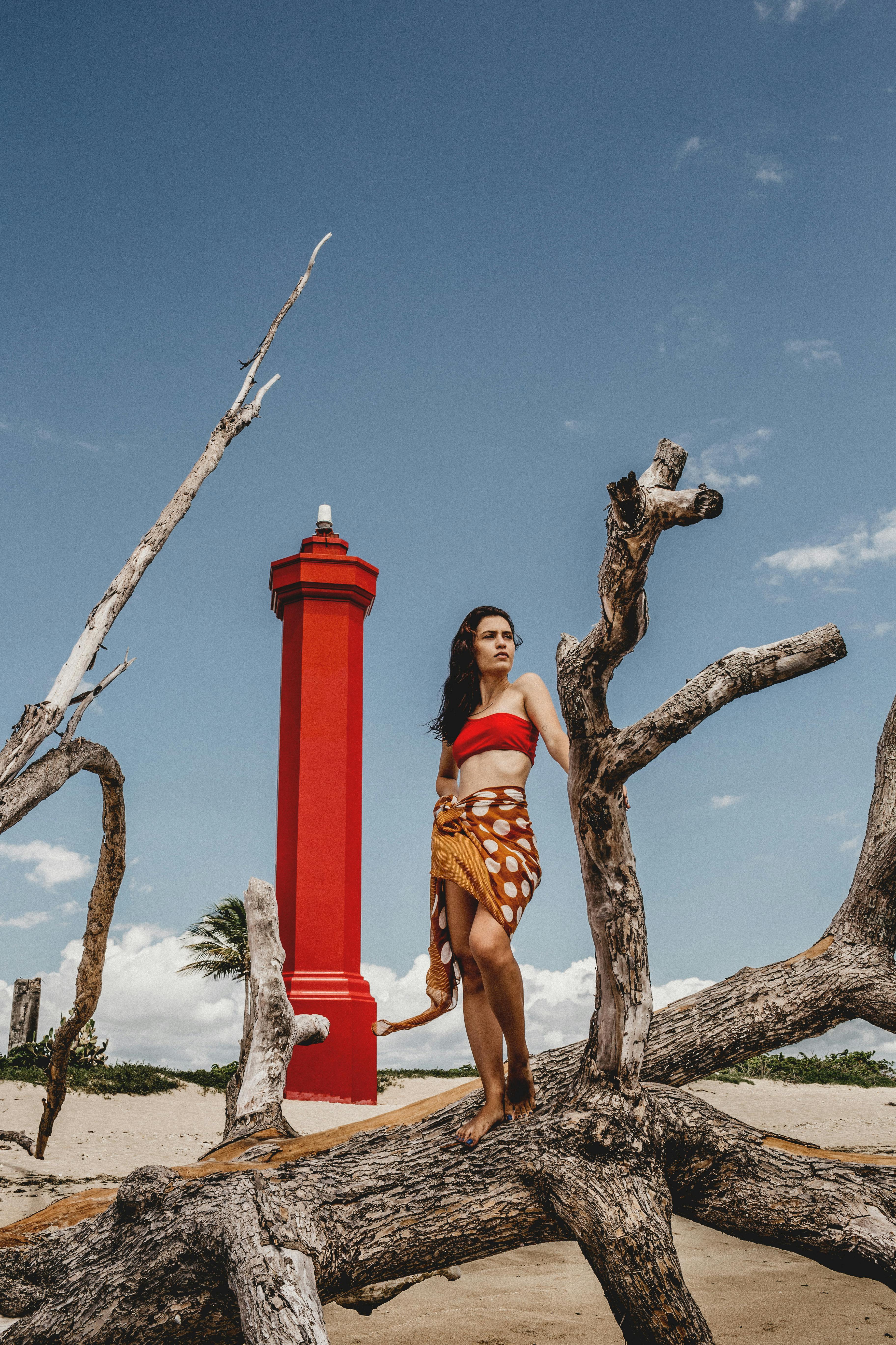 Full body of young female in swimwear and pareo on dry tree of sandy shore with tall red lighthouse