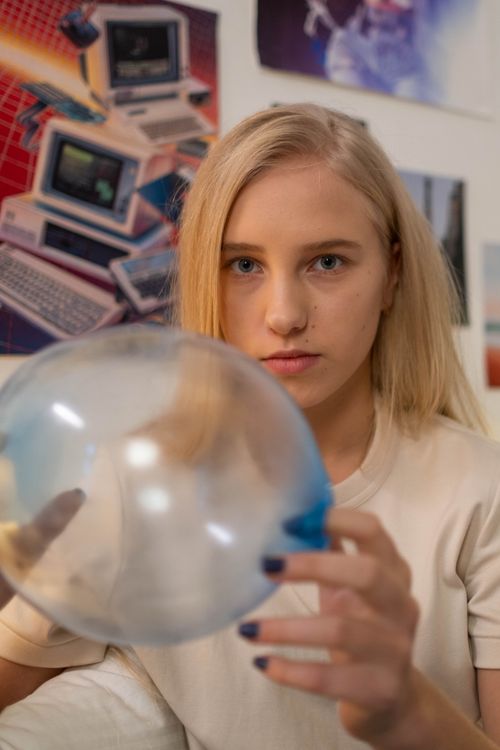 Free Close-up of a blonde woman holding a transparent balloon indoors, with retro computer posters in the background. Stock Photo