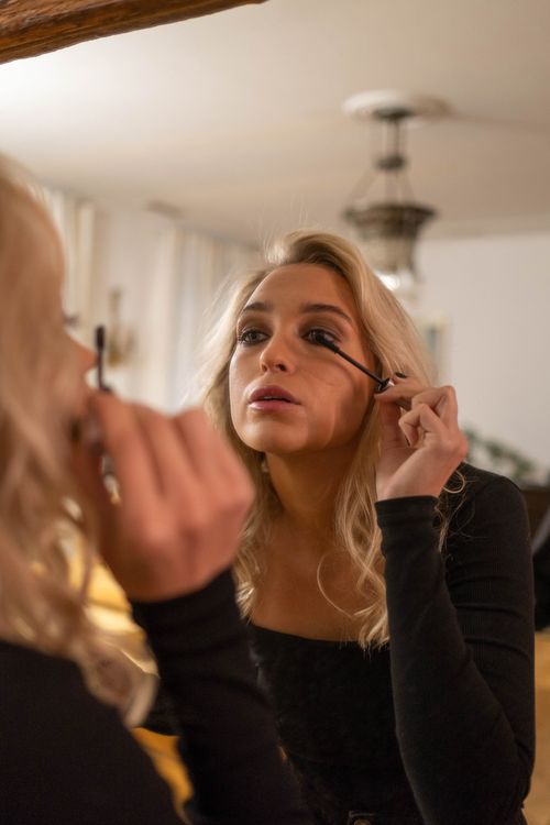 Free A young blonde woman applies mascara while looking into a mirror indoors, capturing her reflection. Stock Photo