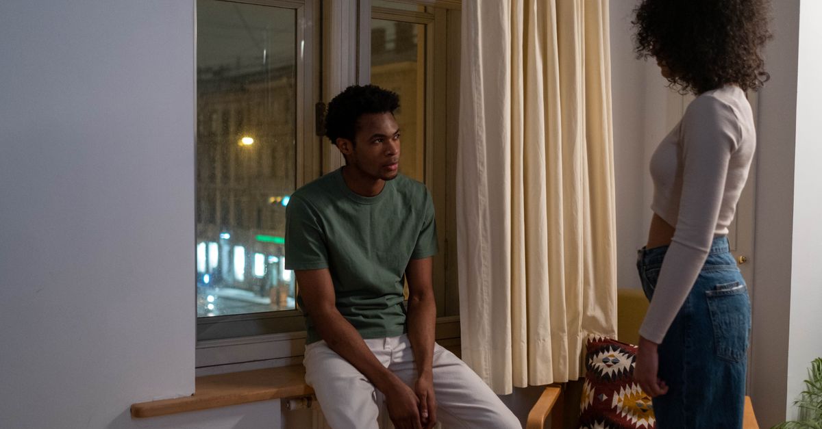 A couple engaged in a tense conversation by a windowsill in a cozy living room setting.