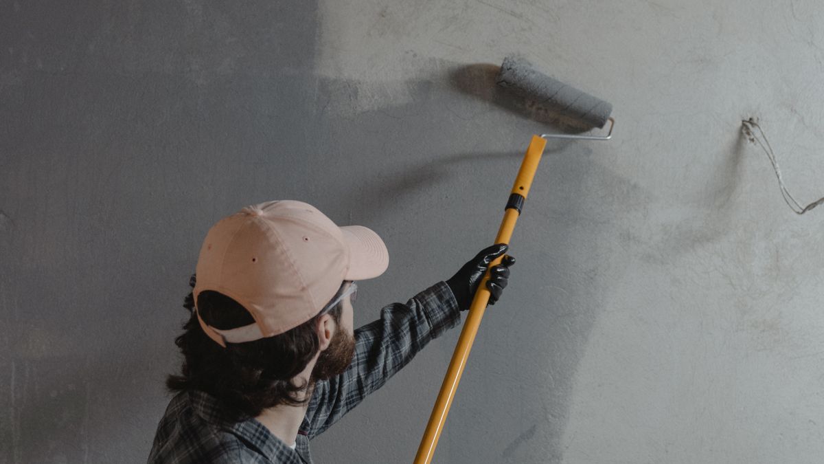 Pintor de boné usando um rolo com cabo longo para pintar uma parede de cinza