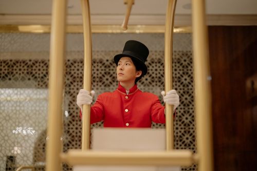 Free A bellboy in a red uniform stands with a luggage cart inside a luxurious hotel. Stock Photo