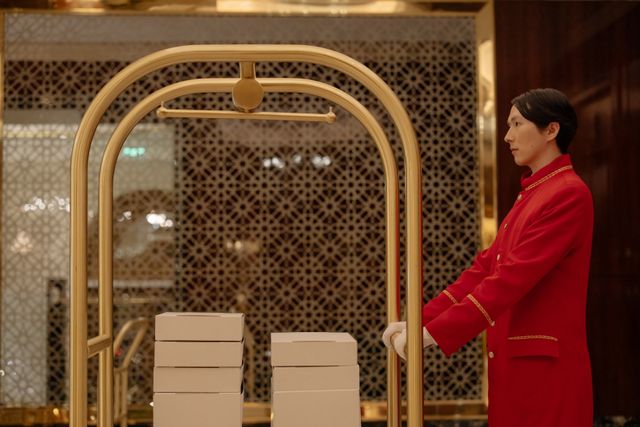 A hotel bellboy in uniform pushes a luggage cart with boxes indoors, demonstrating hospitality service. Related image for Hotel Operations: How Brands Can Improve Performance.