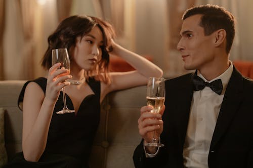 Man in Black Suit Holding Clear Drinking Glass Beside Woman in Black Sleeveless Dress