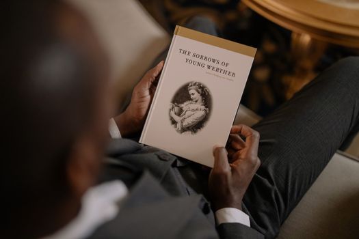 A man in a suit reads "The Sorrows of Young Werther" in an elegant setting.