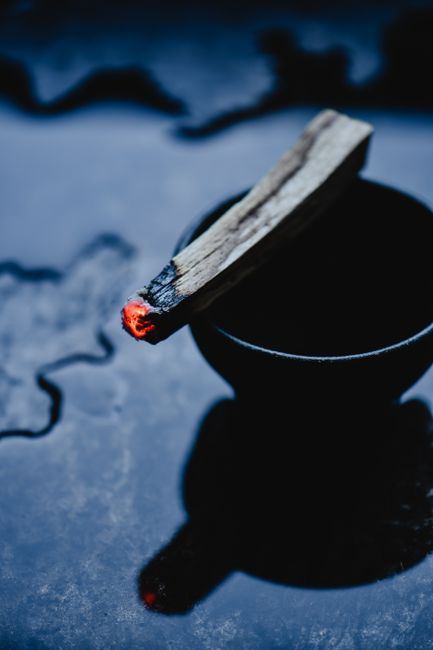 A glowing Palo Santo stick rests on a black bowl, emitting smoke in a ritual setting.