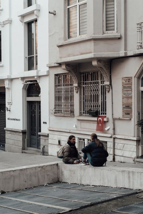 Free Women in outerwear sitting on stone parapet in city street and having conversation near old building with doors and windows while looking at each other Stock Photo