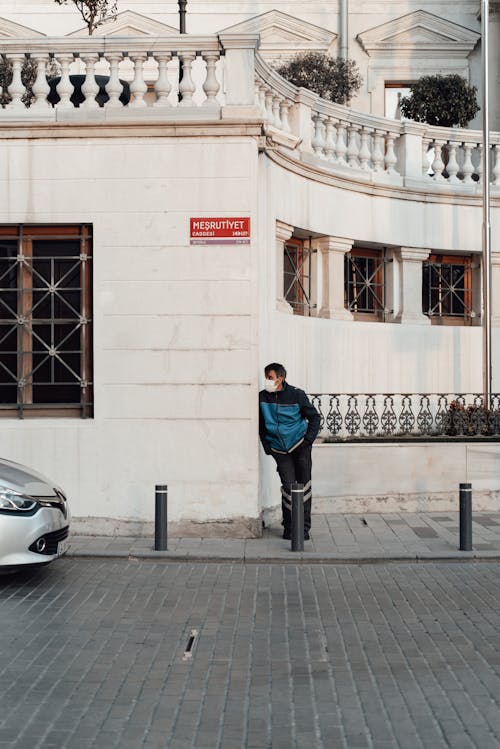 Full body of male in casual clothes and mask leaning on building wall on sidewalk near pillars and road with car in town district in daytime while looking away