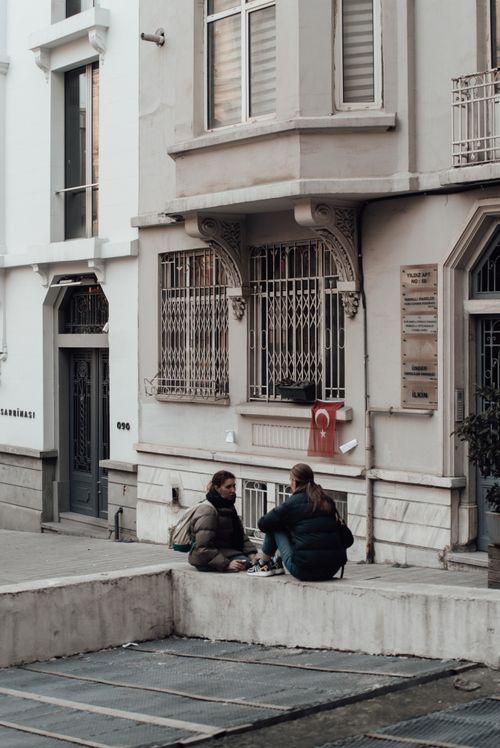 Free Ladies in warm clothes sitting on stone border while having conversation near aged building with doors and windows in city street in daytime while looking at each other Stock Photo