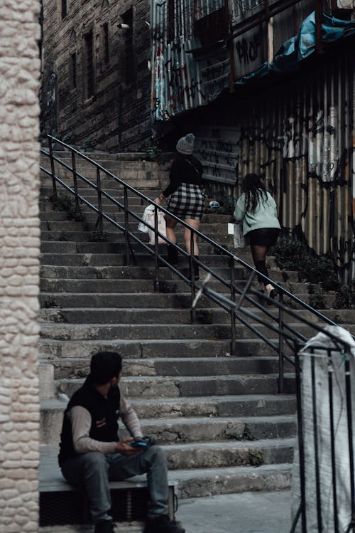 Full body of anonymous women on stone stairway with railing near man sitting and watching at ladies near buildings in city street in daylight