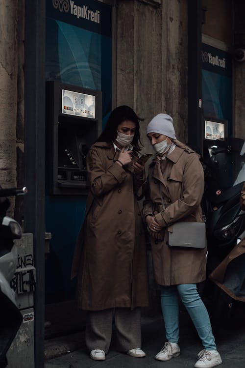 Full body of female friends in outerwear and masks standing on pavement in town street while using cellphone near ATMs in daytime