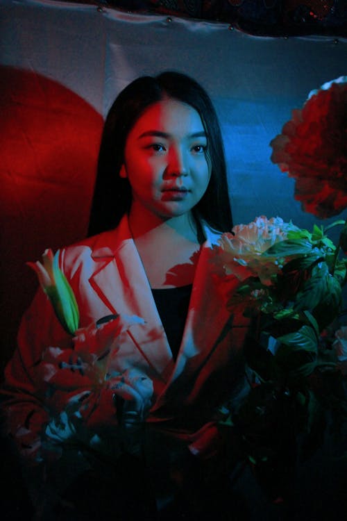 Free Serious Asian female with long hair wearing white jacket standing with blossoming flowers in dark place Stock Photo