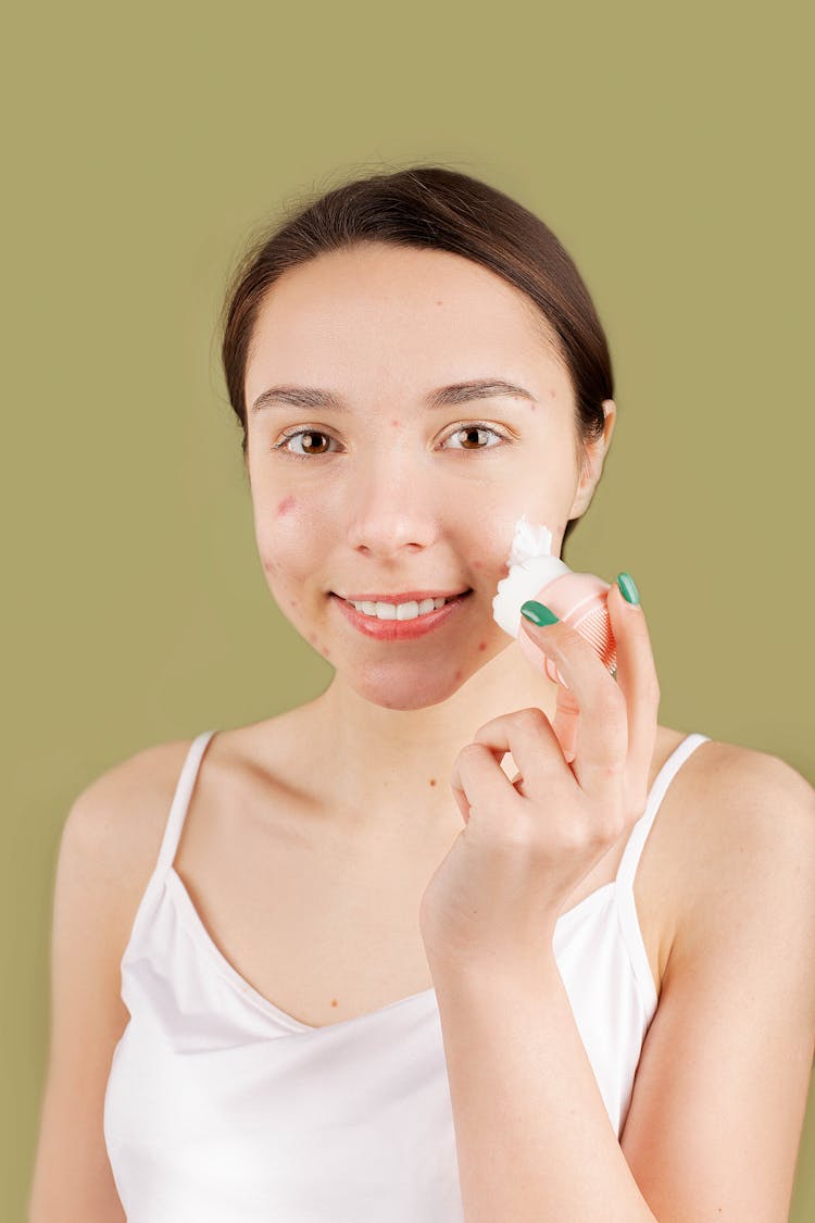 Smiling Teenager Applying A Facial Cream On Her Face While Looking At Camera