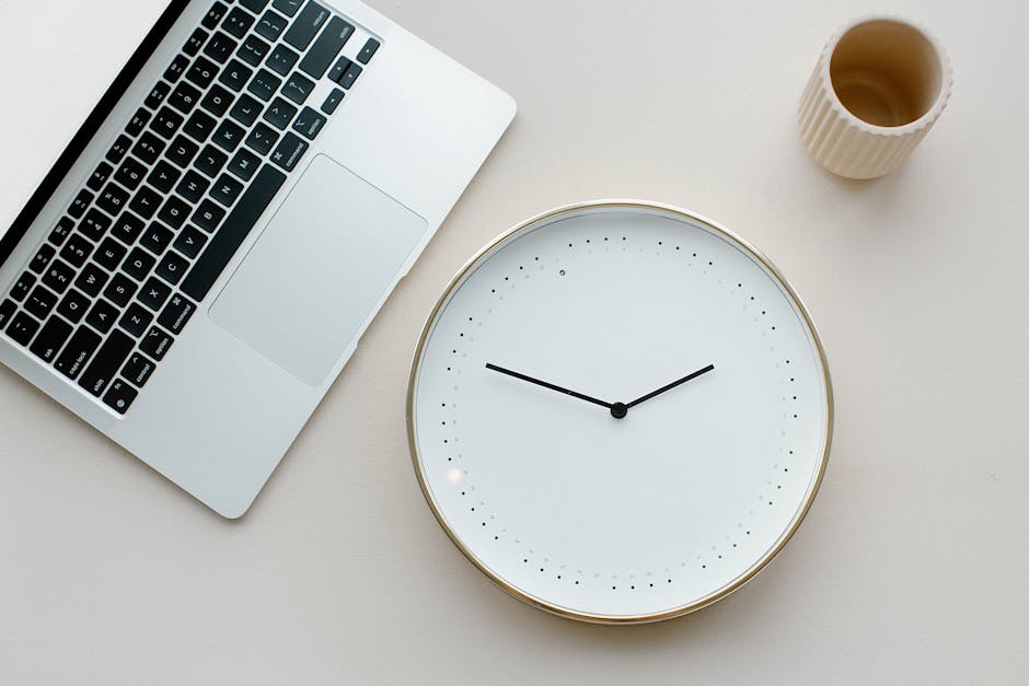 A clean minimalist workspace featuring a laptop, wall clock, and cup on a neutral background.
