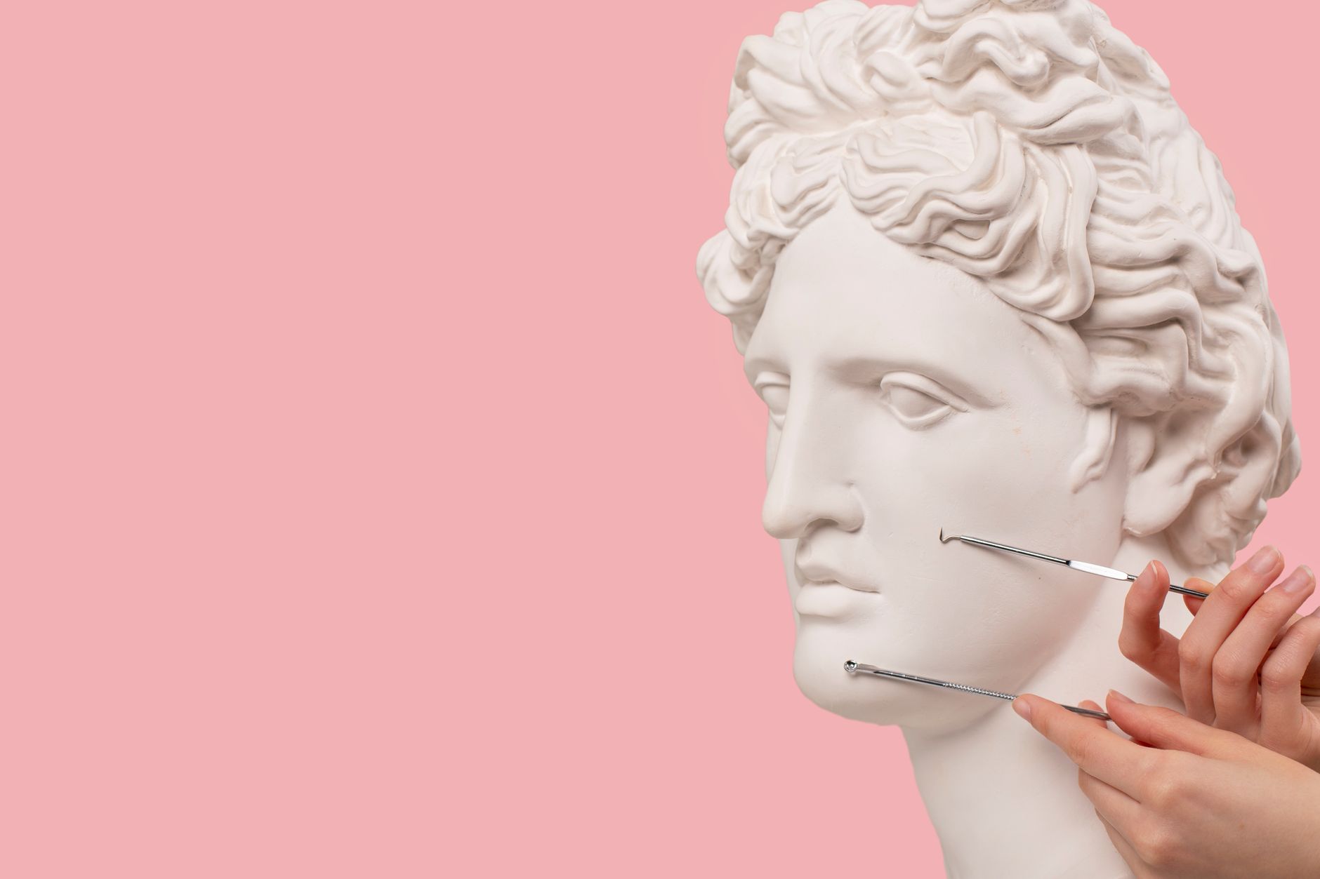 Classical bust with beauty tools on pink background, merging art with skincare.