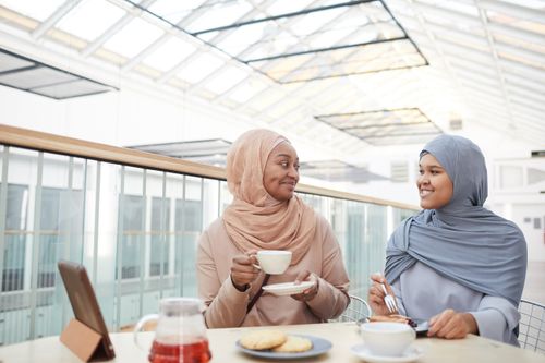 Free Two women sharing coffee and pastries in a bright indoor setting, wearing hijabs and smiling. Stock Photo