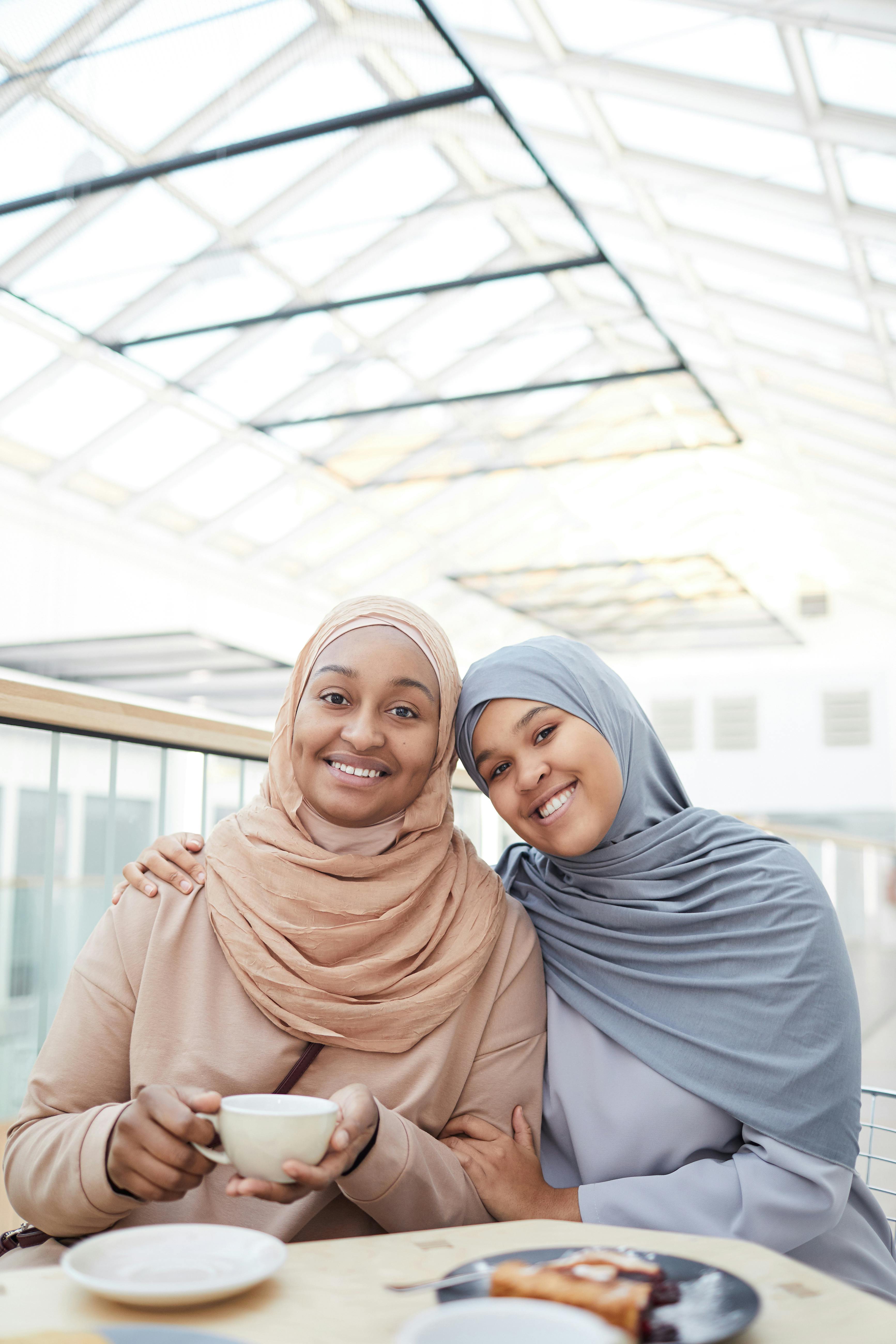 Two women in hijabs smiling and enjoying coffee indoors under natural lighting.