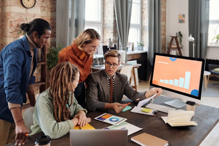 A Group Of People Discussing Beside A Desktop With Graph On Screen