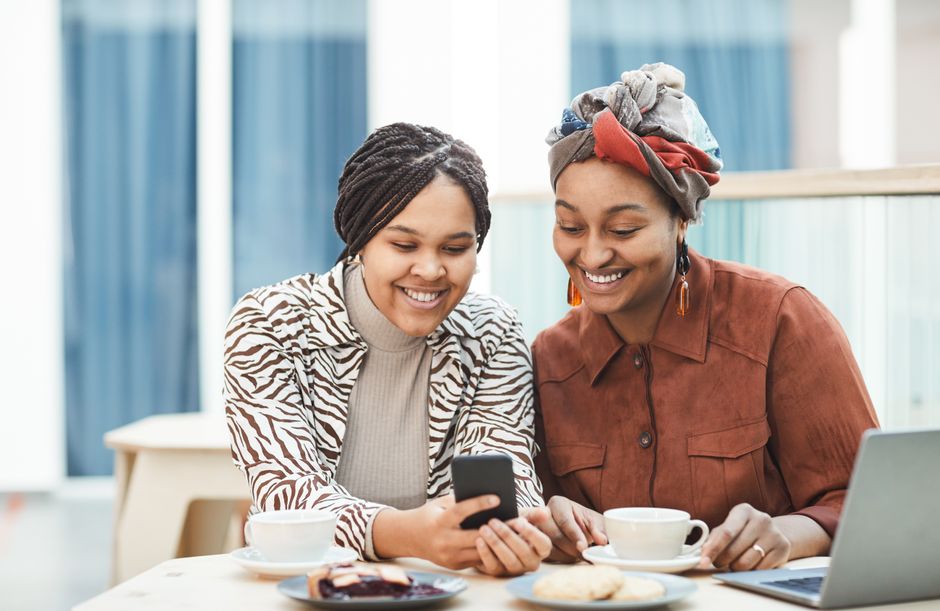 Two African American women with braided hairstyles enjoy coffee and smartphone interaction at a café.