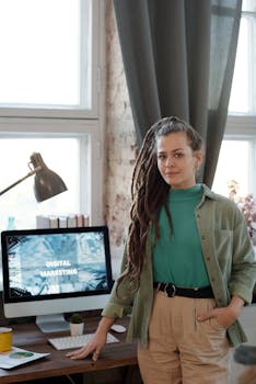 Confident young woman standing in a modern home office with a computer screen displaying digital marketing.