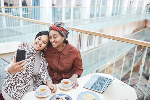 Free Two women in a stylish cafe enjoying tea and capturing a selfie with a smartphone. Stock Photo